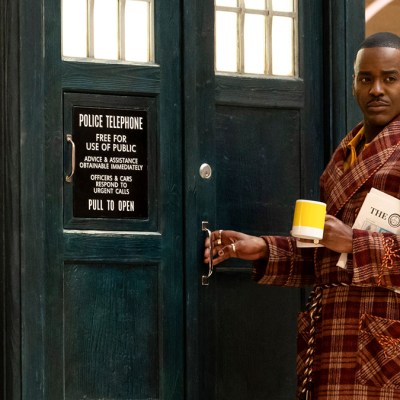 Ncuti Gatwa wearing a tartan dressing gown and holding a cup of tea standing by the TARDIS